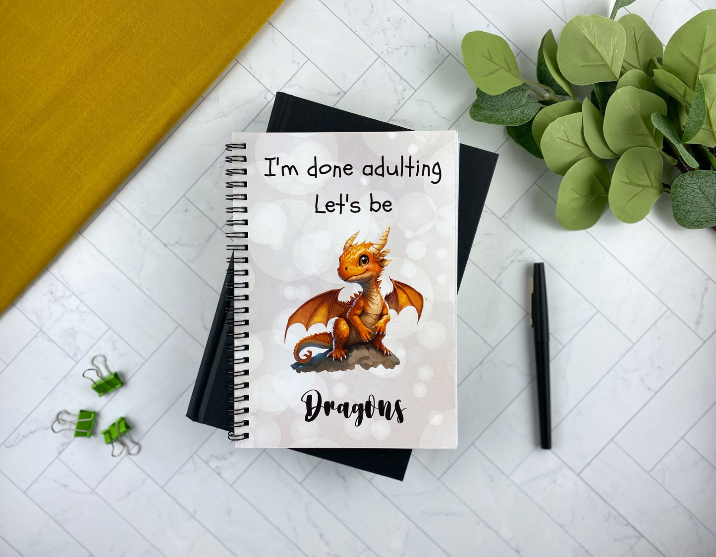 Spiral notebook cover with a soft bokeh background, an orange baby dragon perched on a rock, and the words "I'm done adulting. Let's be dragons," resting on a black notebook with green binder clips, a black pen, and eucalyptus greenery nearby.