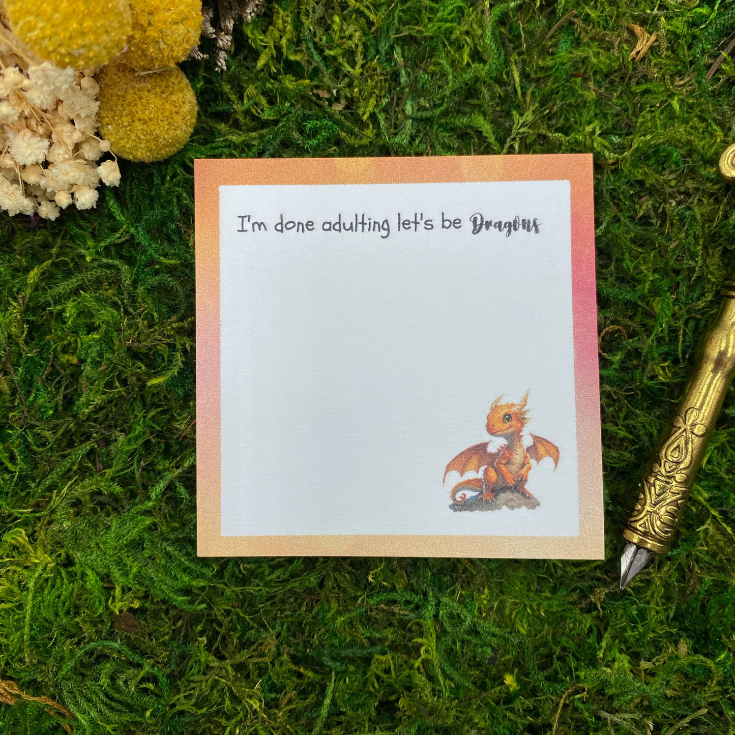 Square sticky note pad with an orange-to-white gradient border, an orange baby dragon perched on a rock, and the words "I'm done adulting, let's be dragons," resting on moss next to dried yellow and white flowers and a gold quill pen.