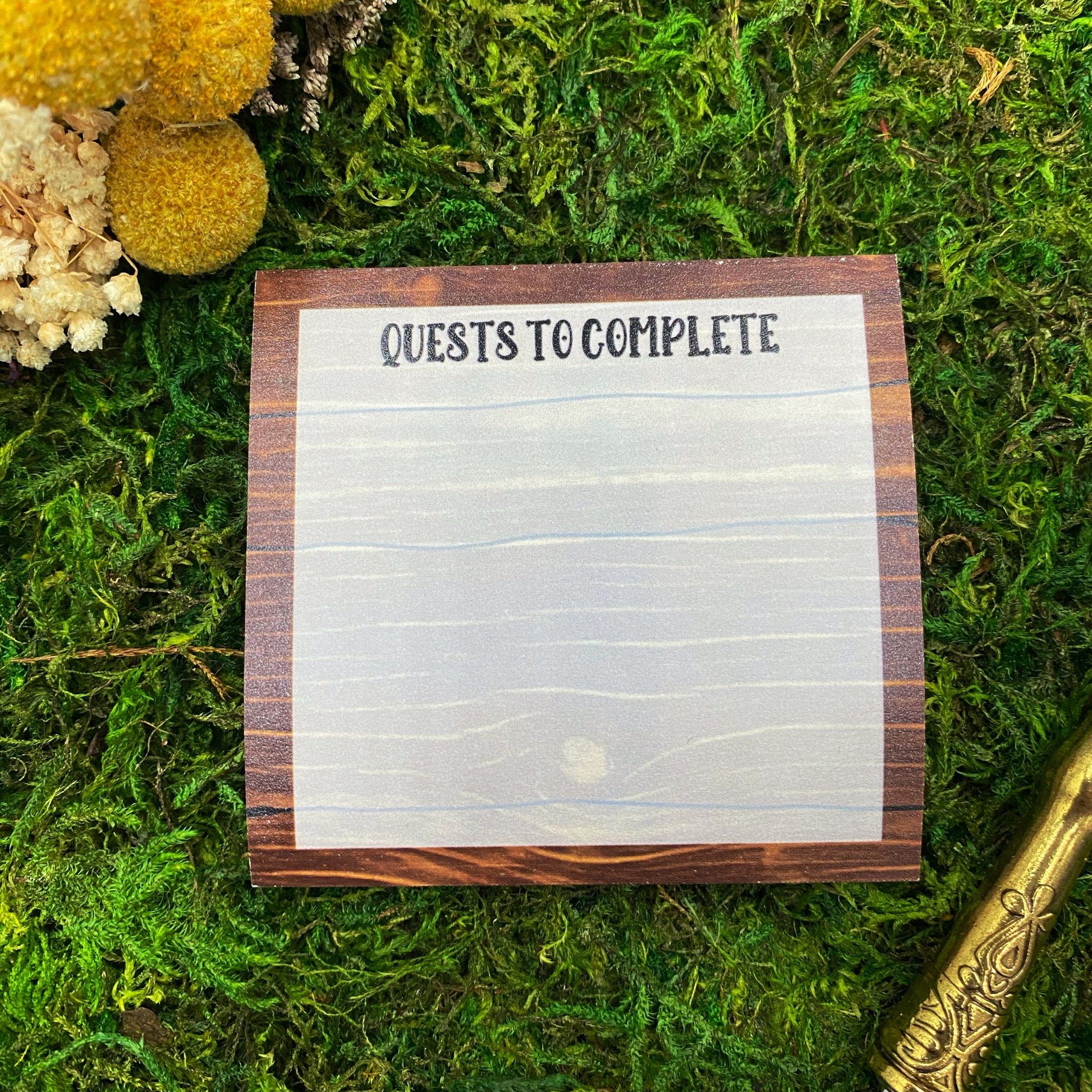 Square sticky note pad with a wood-plank border designed to look like a tavern quest board, headed "Quests to Complete," resting on moss next to dried yellow and white flowers and a gold quill pen.