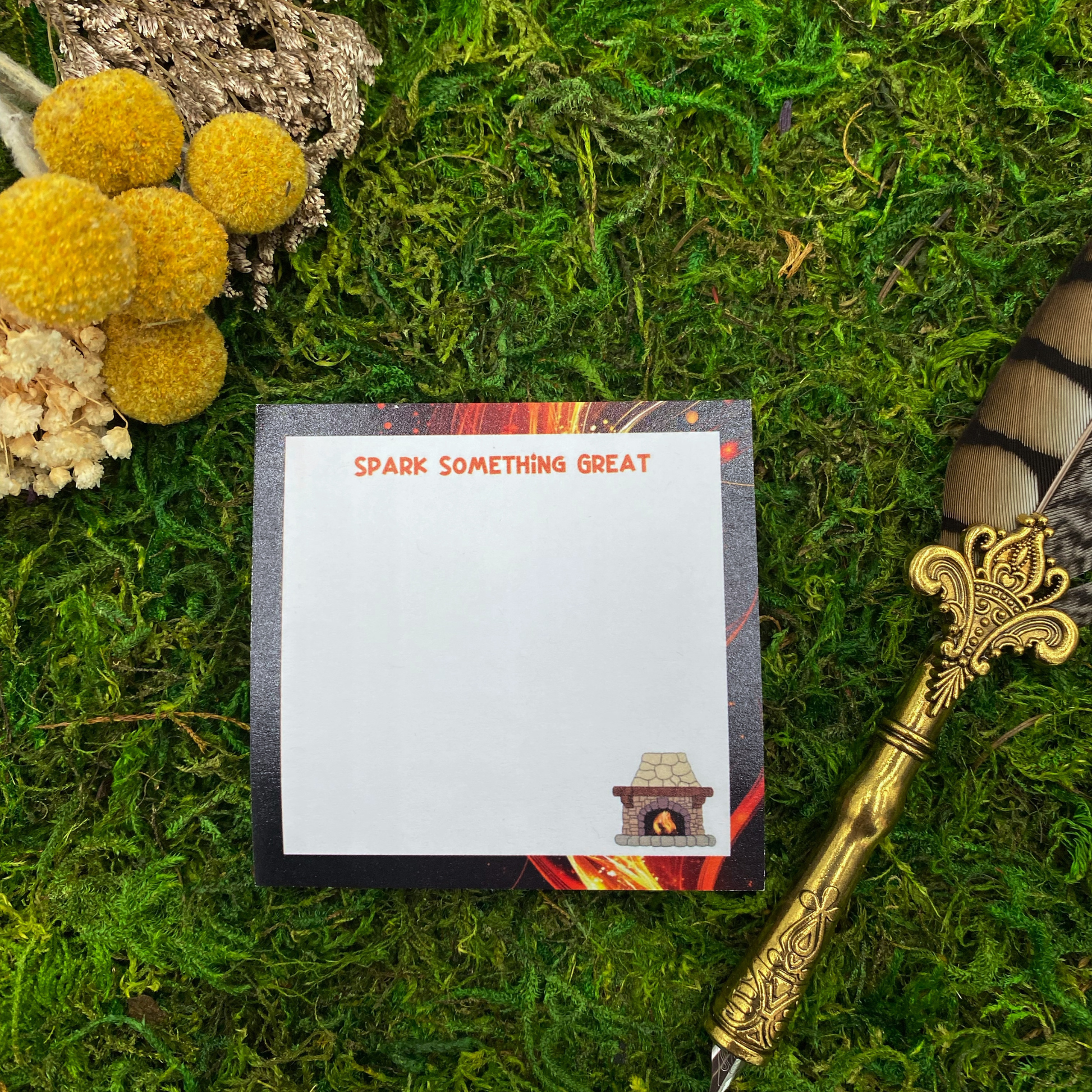 Square sticky note pad with a dark fiery-streaked border, a small illustrated stone fireplace, and the words "Spark Something Great," resting on moss next to dried yellow and white flowers and a gold quill pen.