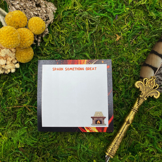 Square sticky note pad with a dark fiery-streaked border, a small illustrated stone fireplace, and the words "Spark Something Great," resting on moss next to dried yellow and white flowers and a gold quill pen.