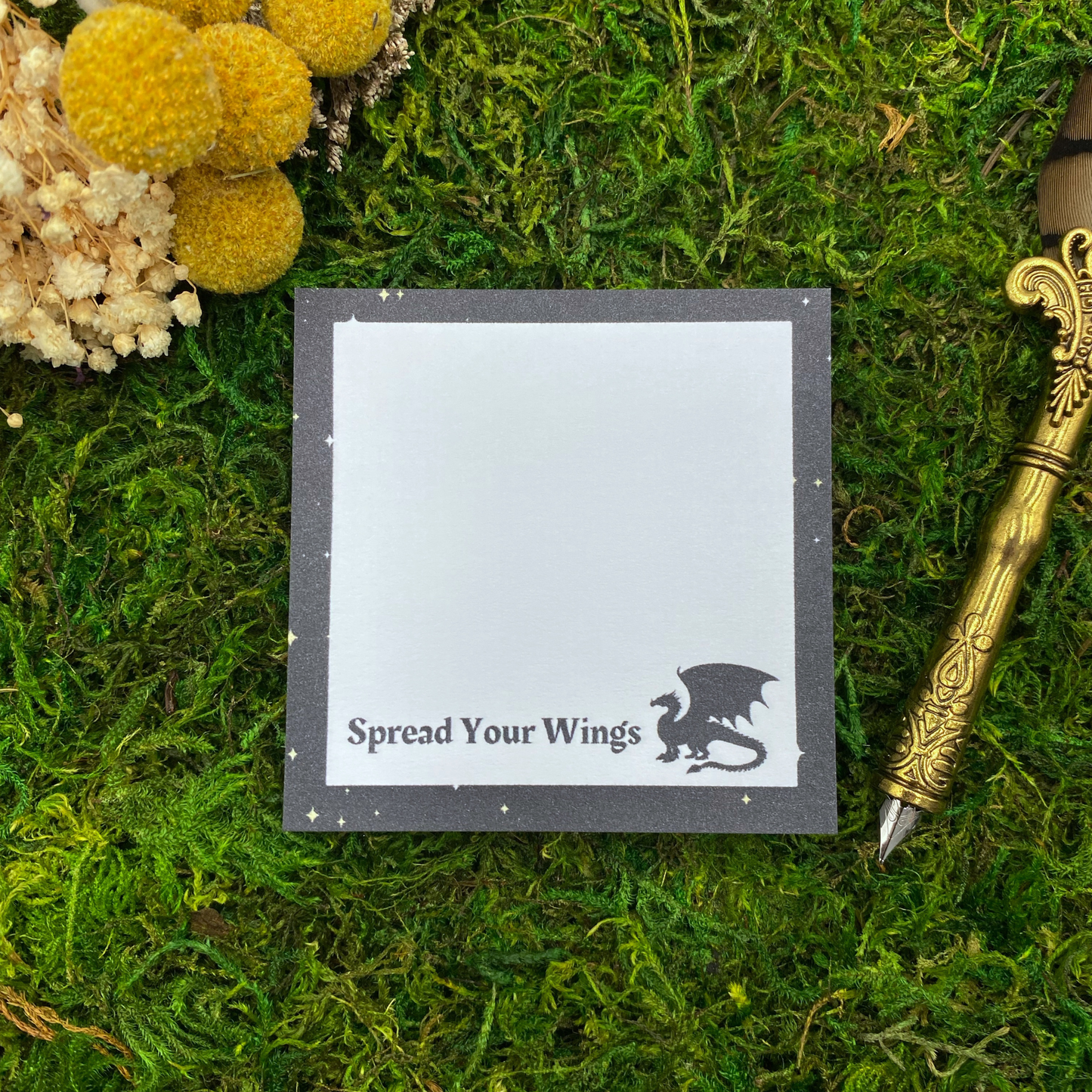 Square sticky note pad with a dark starry border, a black dragon silhouette, and the words "Spread Your Wings," resting on moss next to dried yellow and white flowers and a gold quill pen.