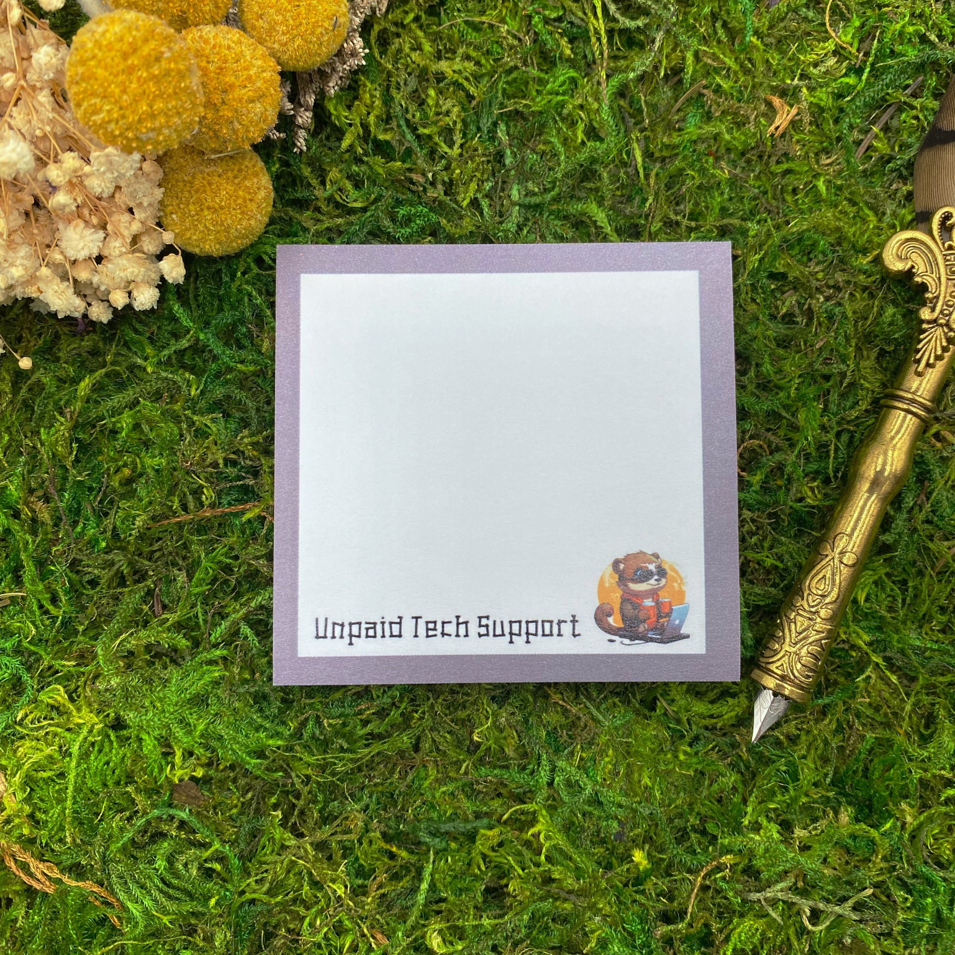 Square sticky note pad with a lavender border, an illustrated otter wearing sunglasses at a laptop, and the words "Unpaid Tech Support," resting on moss next to dried yellow and white flowers and a gold quill pen.