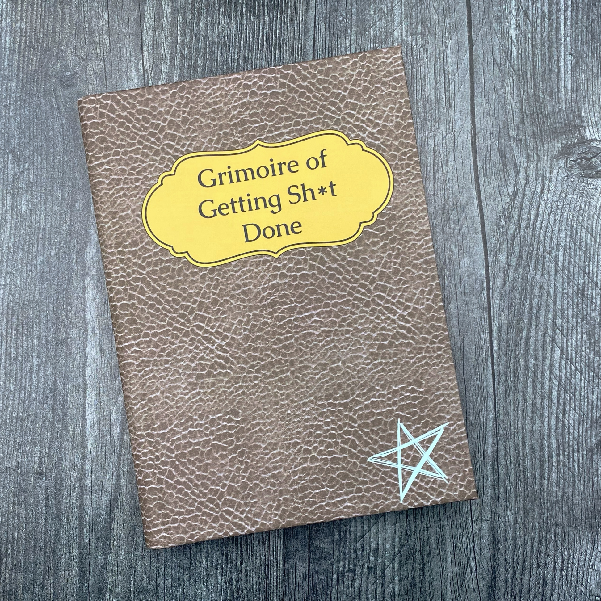 Journal cover with a brown textured leather-look design, a yellow label reading "Grimoire of Getting Sh*t Done," and a hand-drawn pentagram, on a wood surface.