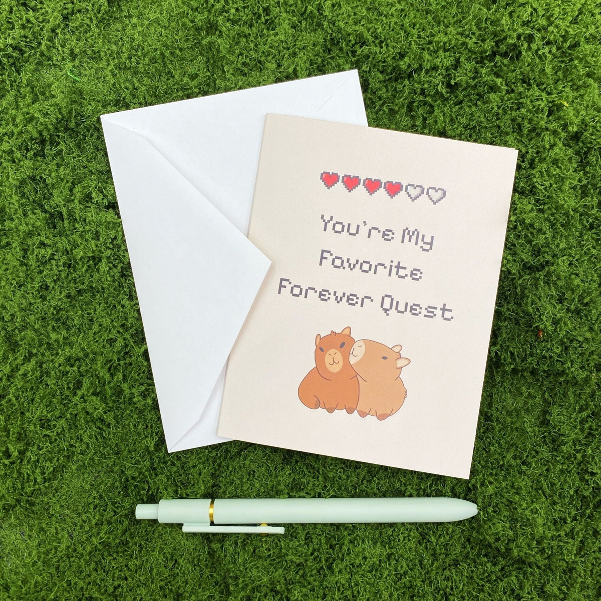"You're My Favorite Forever Quest" greeting card lying on a white envelope on grass, with a mint green pen beside it for scale.