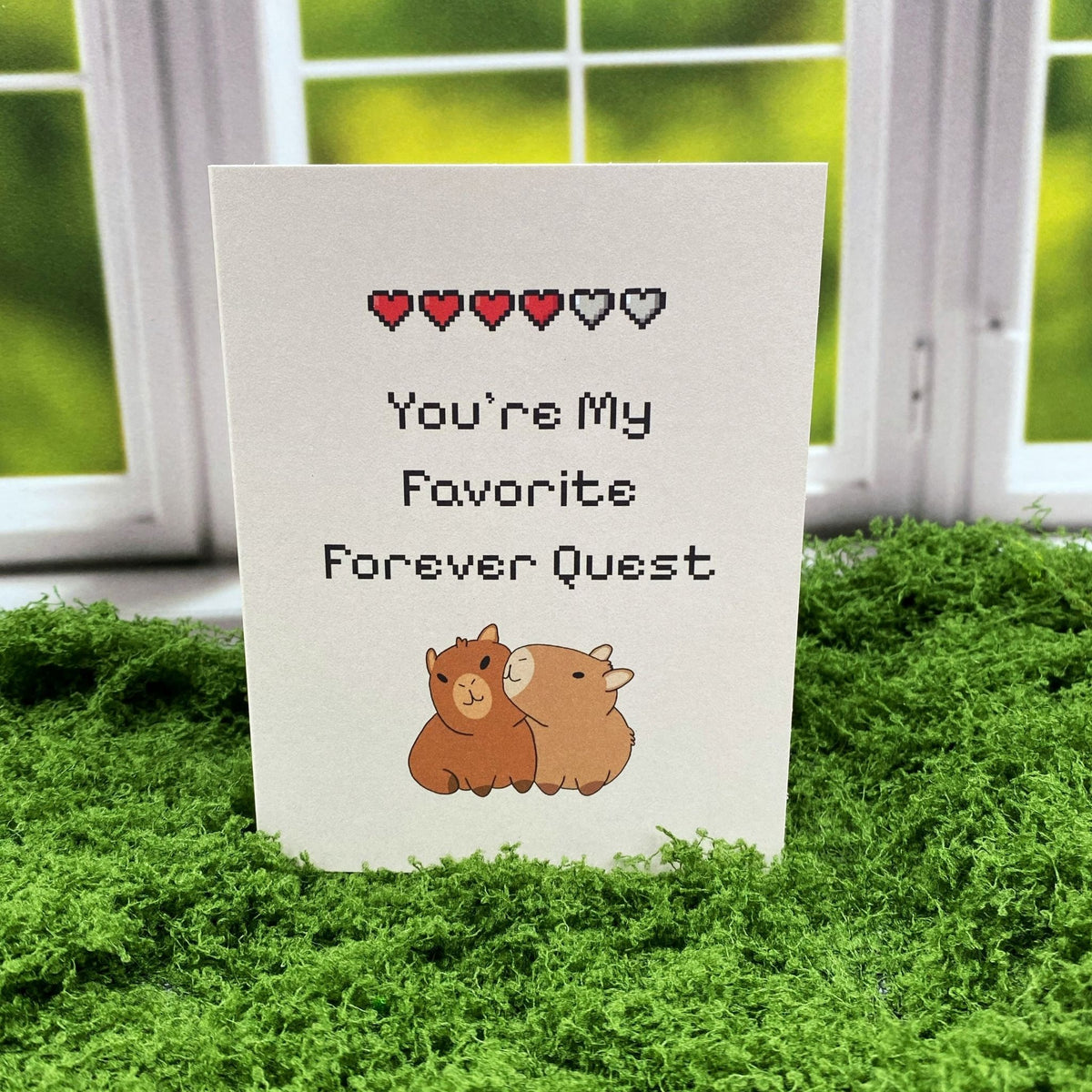 "You're My Favorite Forever Quest" greeting card standing upright on a moss mat in front of a window backdrop, featuring a pixel-art heart health bar and two illustrated capybaras cuddling.