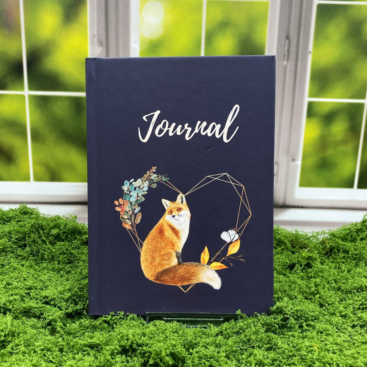 Navy hardcover journal reading 'Journal' with a red fox illustration inside a geometric heart wreath of leaves and a butterfly, displayed on a moss mat with a window backdrop.