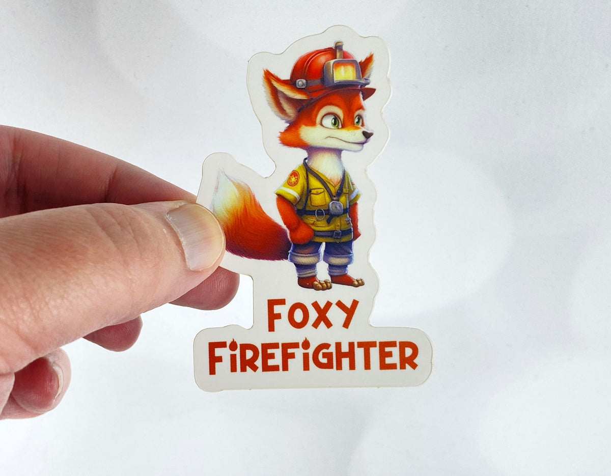 A hand holds a sticker illustration of a fox dressed in firefighter gear with a helmet, reading "Foxy Firefighter," against a white background.
