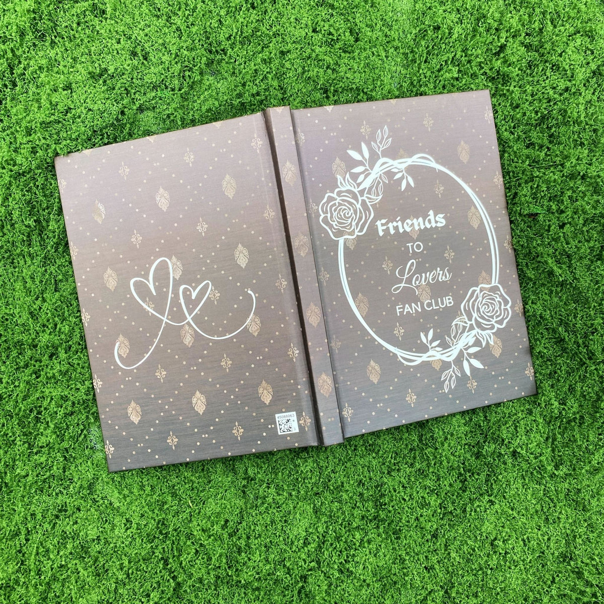 Open brown hardcover journal showing two intertwined heart outlines on the back cover and the rose wreath 'Friends to Lovers Fan Club' design on the front cover.