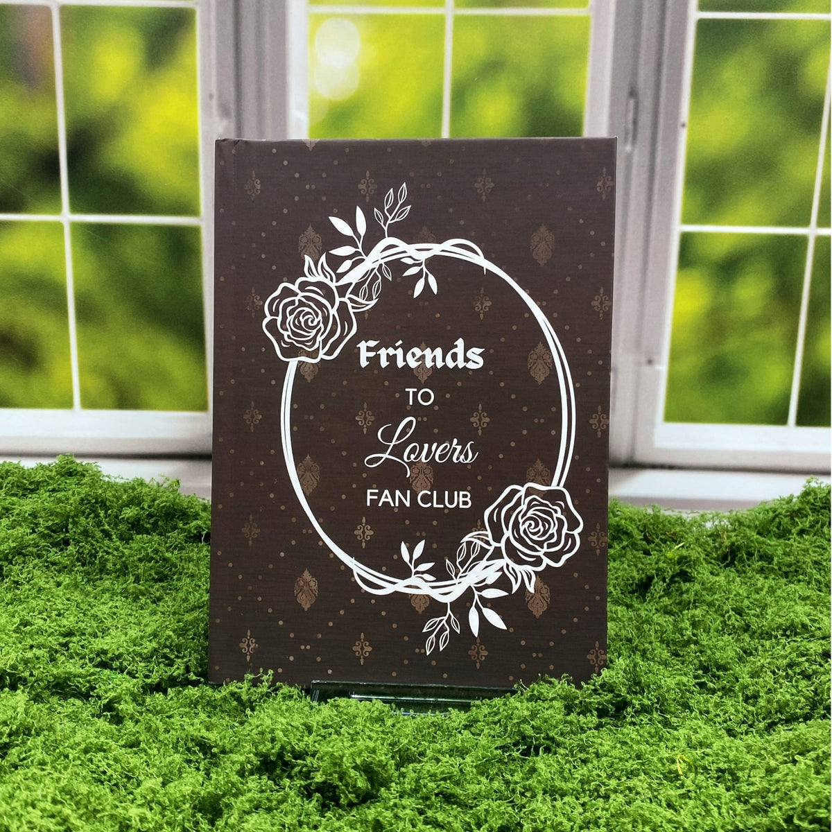 Brown hardcover journal with a damask pattern and a rose wreath frame around text reading 'Friends to Lovers Fan Club,' displayed on a moss mat with a window backdrop.