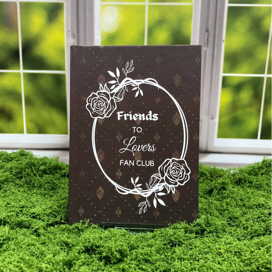 Brown hardcover journal with a damask pattern and a rose wreath frame around text reading 'Friends to Lovers Fan Club,' displayed on a moss mat with a window backdrop.