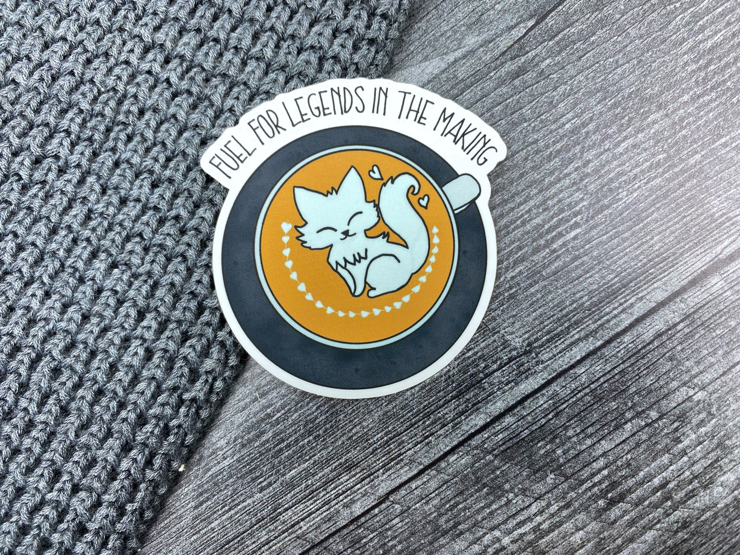 Die-cut vinyl sticker showing a latte with fox and heart foam art, with the text "Fuel for legends in the making," on a gray knit and wood background