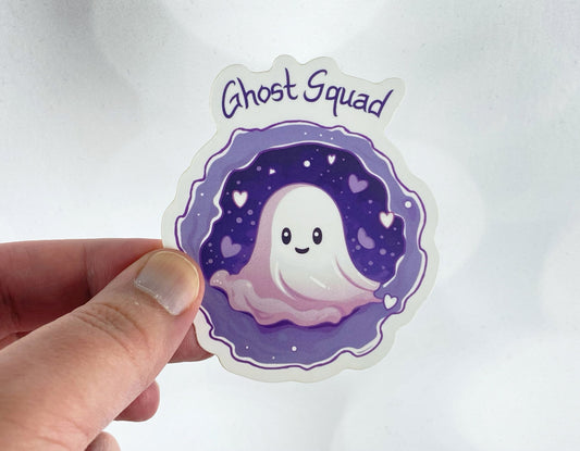 A hand holds a sticker illustration of a smiling ghost surrounded by hearts and stars against a purple background, reading "Ghost Squad," against a white backdrop.