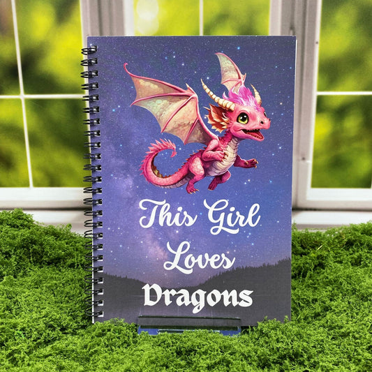 Spiral-bound notebook with a pink dragon flying against a starry night sky, reading 'This Girl Loves Dragons' above a silhouetted mountain landscape, displayed on a moss mat with a window backdrop