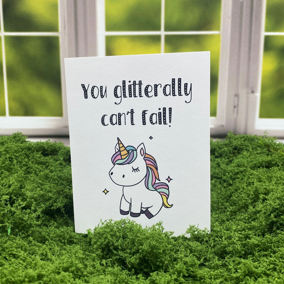 "You Glitterally Can't Fail!" greeting card standing upright on a moss mat in front of a window backdrop, featuring an illustrated unicorn with a pastel rainbow mane.