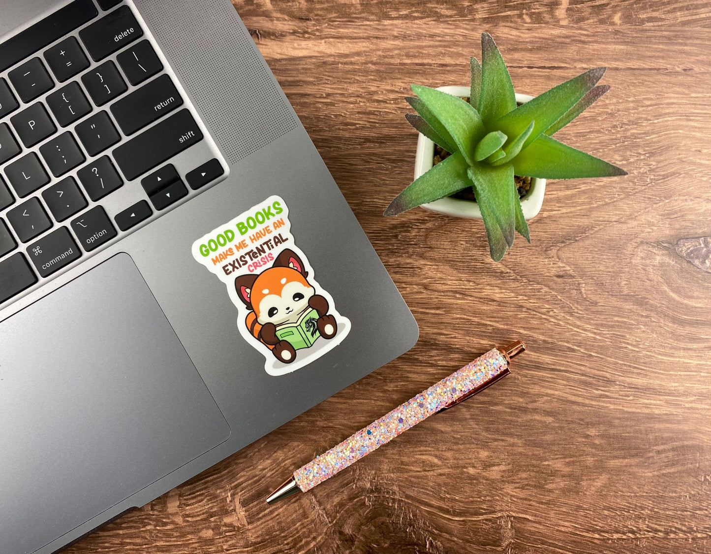 Red panda reading a book sticker applied to the corner of a silver laptop next to a succulent and a glitter pen on a wood desk