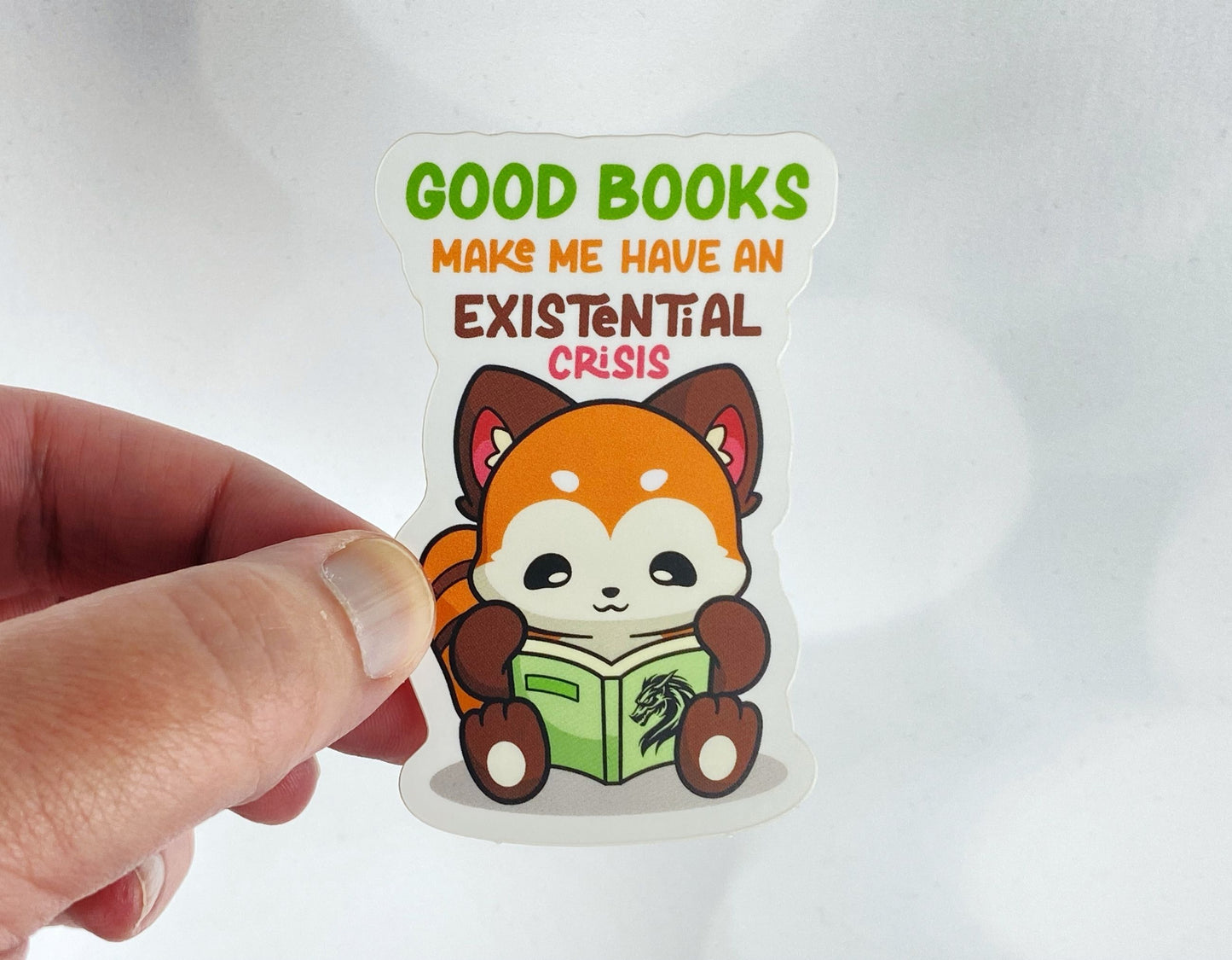 Die-cut vinyl sticker held between fingers, showing a red panda reading a green book with a dragon on the cover, above the text "Good books make me have an existential crisis," on a white background