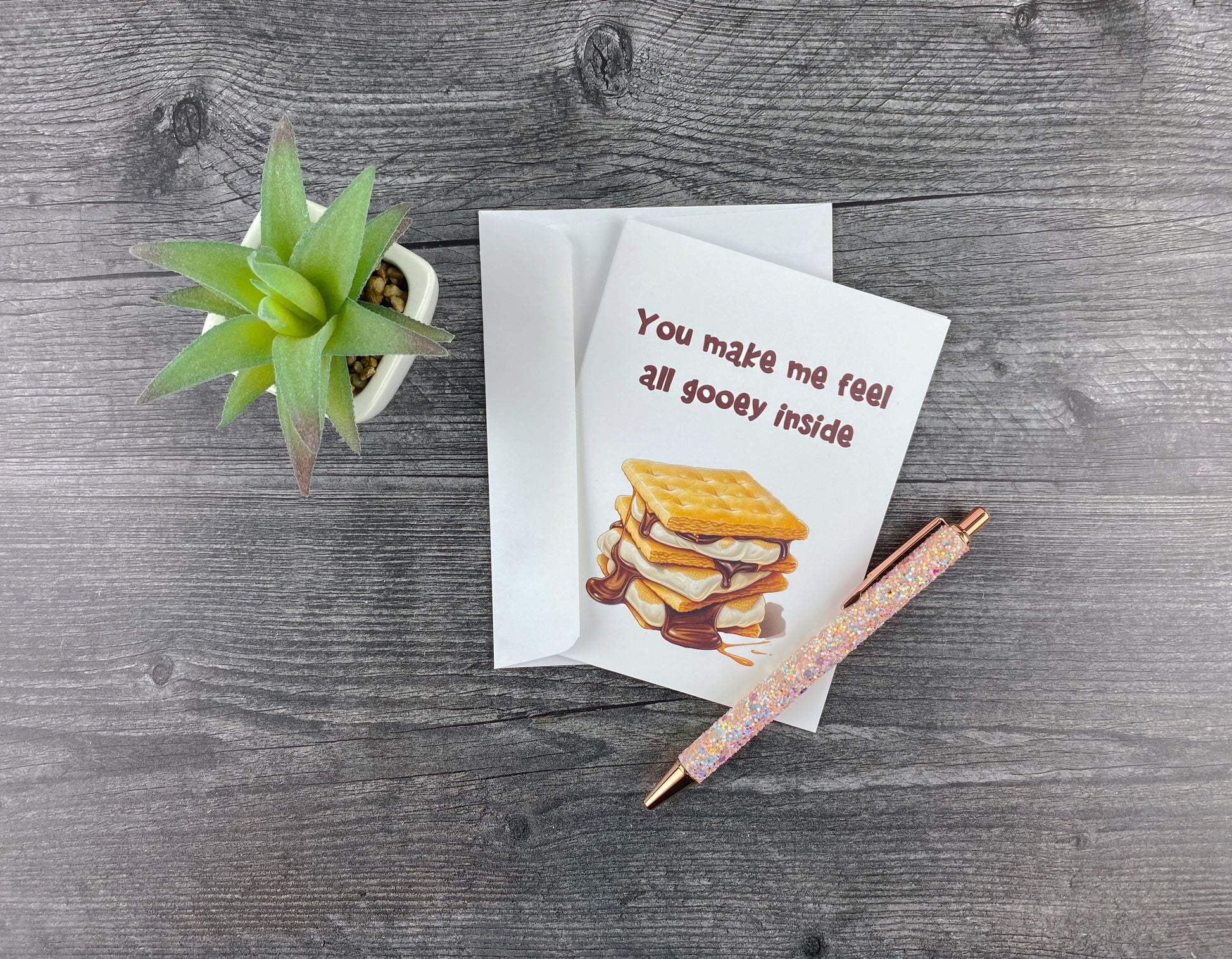 Greeting card and matching envelope, front featuring an illustrated dripping s'more and reading "You make me feel all gooey inside," resting on a grey wood surface next to a pink glitter pen and small succulent.