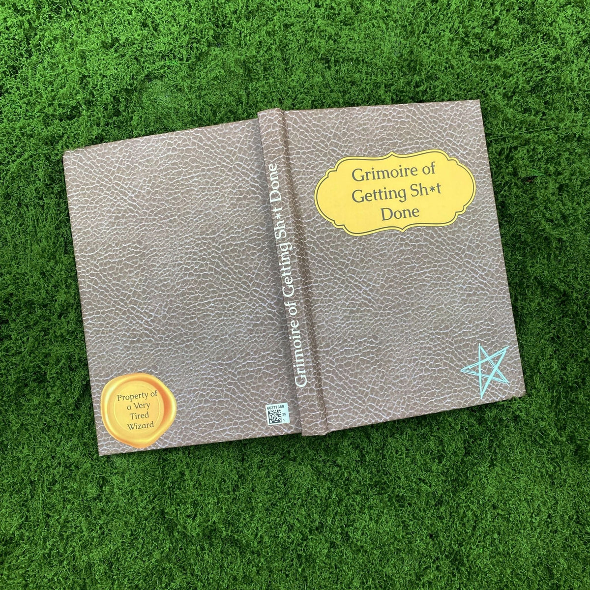 Open faux-leather hardcover journal showing a gold wax-seal-style stamp reading 'Property of a Very Tired Wizard' on the inside back cover and the 'Grimoire' title on the front cover.