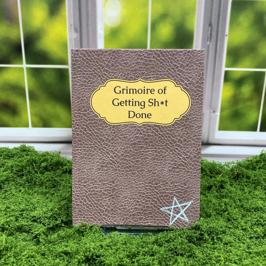 Faux-leather hardcover journal with a pentagram accent, reading 'Grimoire of Getting Sh*t Done' on a yellow label, displayed on a moss mat with a window backdrop