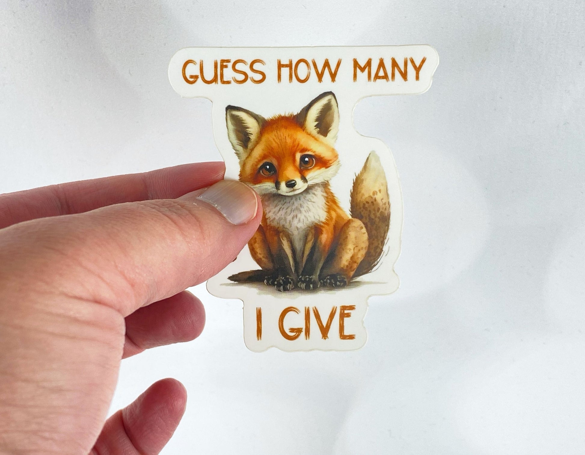 A hand holds a sticker illustration of a wide-eyed fox with the text "Guess how many I give" against a white background.