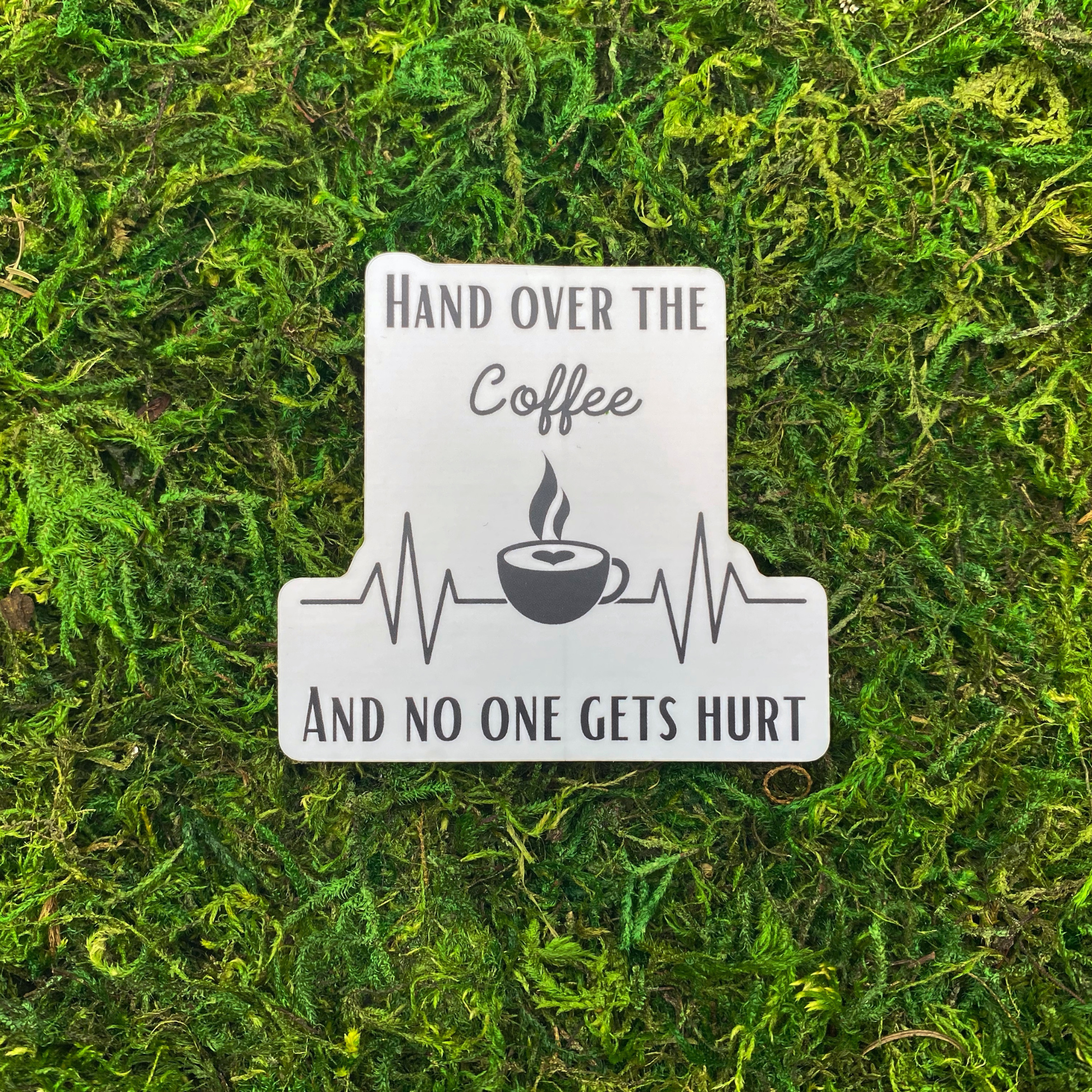 A sticker reading "Hand over the coffee and no one gets hurt" with a heartbeat monitor line and a steaming coffee cup, resting on artificial moss.