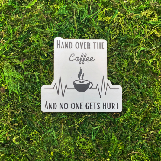 A sticker reading "Hand over the coffee and no one gets hurt" with a heartbeat monitor line and a steaming coffee cup, resting on artificial moss.