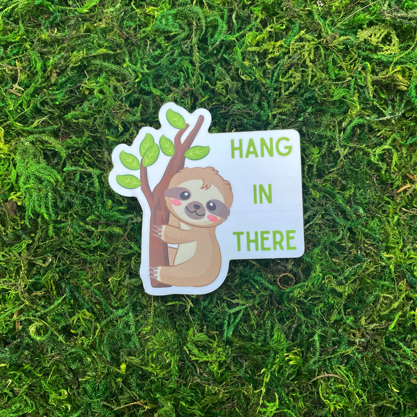 A sticker with an illustration of a cute sloth clinging to a tree branch beside the text "Hang in there," resting on artificial moss.