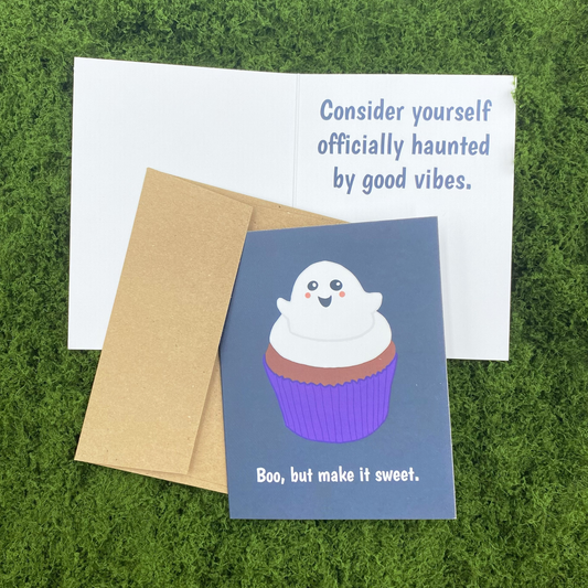 A2 matte Halloween greeting card showing front cover with kawaii ghost cupcake illustration and the text "Boo, but make it sweet," alongside the open interior message "Consider yourself officially haunted by good vibes," with a kraft envelope, on a green moss background.