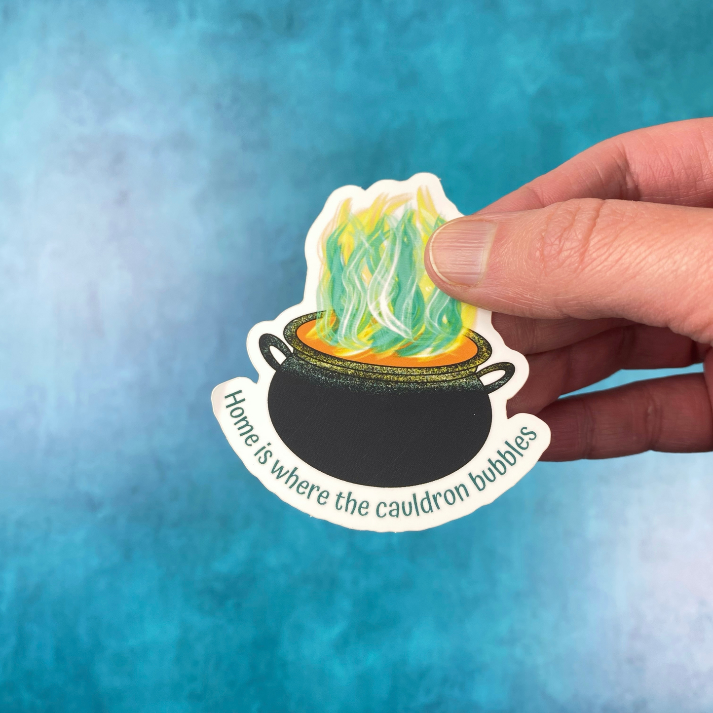 Die-cut vinyl sticker showing a black cauldron with green and yellow steam rising from it, with the text "Home is where the cauldron bubbles," on a teal background