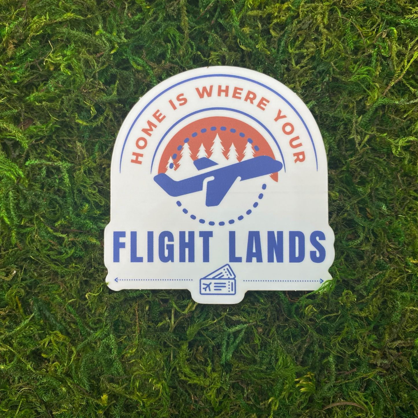 A sticker reading "Home is where your flight lands" with an illustration of an airplane flying in front of a forest skyline, resting on artificial moss.