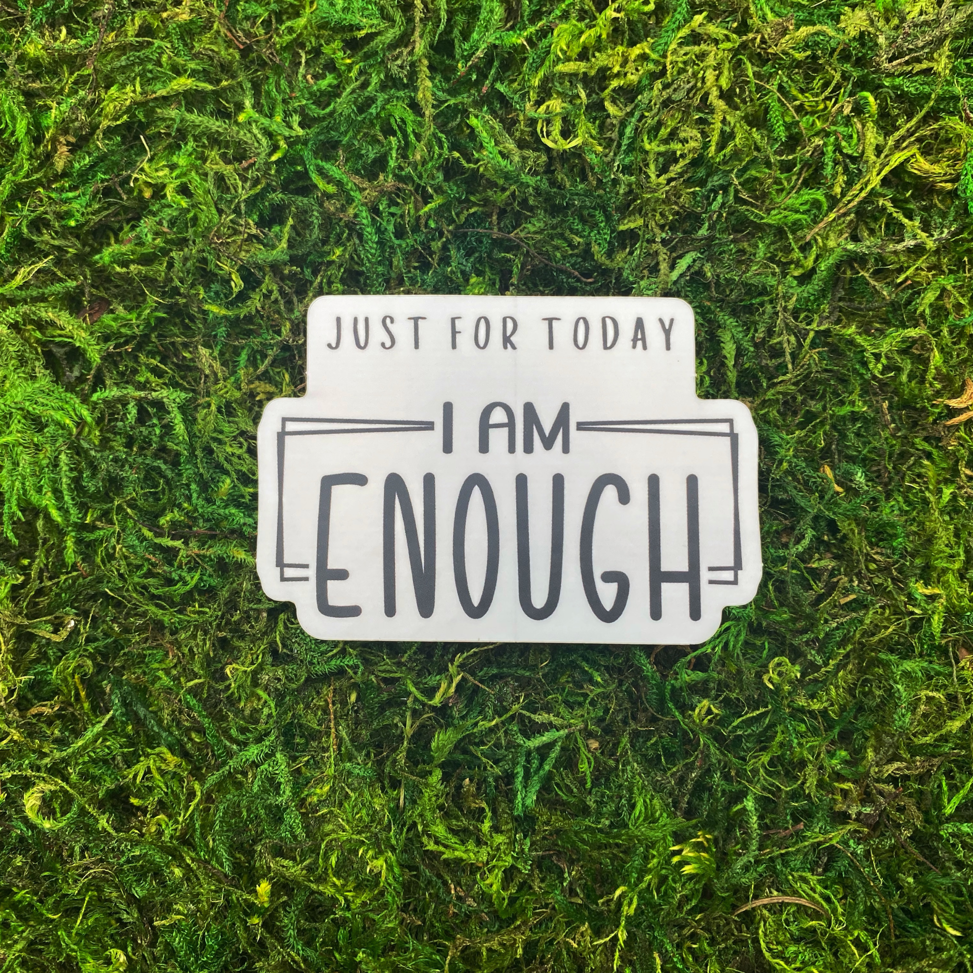 A sticker reading "Just for today, I am enough" in bold hand-lettered text, resting on artificial moss.