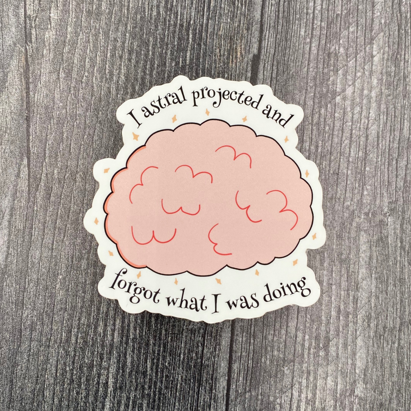Die-cut vinyl sticker showing a pink brain surrounded by sparkles, with the text "I astral projected and forgot what I was doing," on a gray wood background