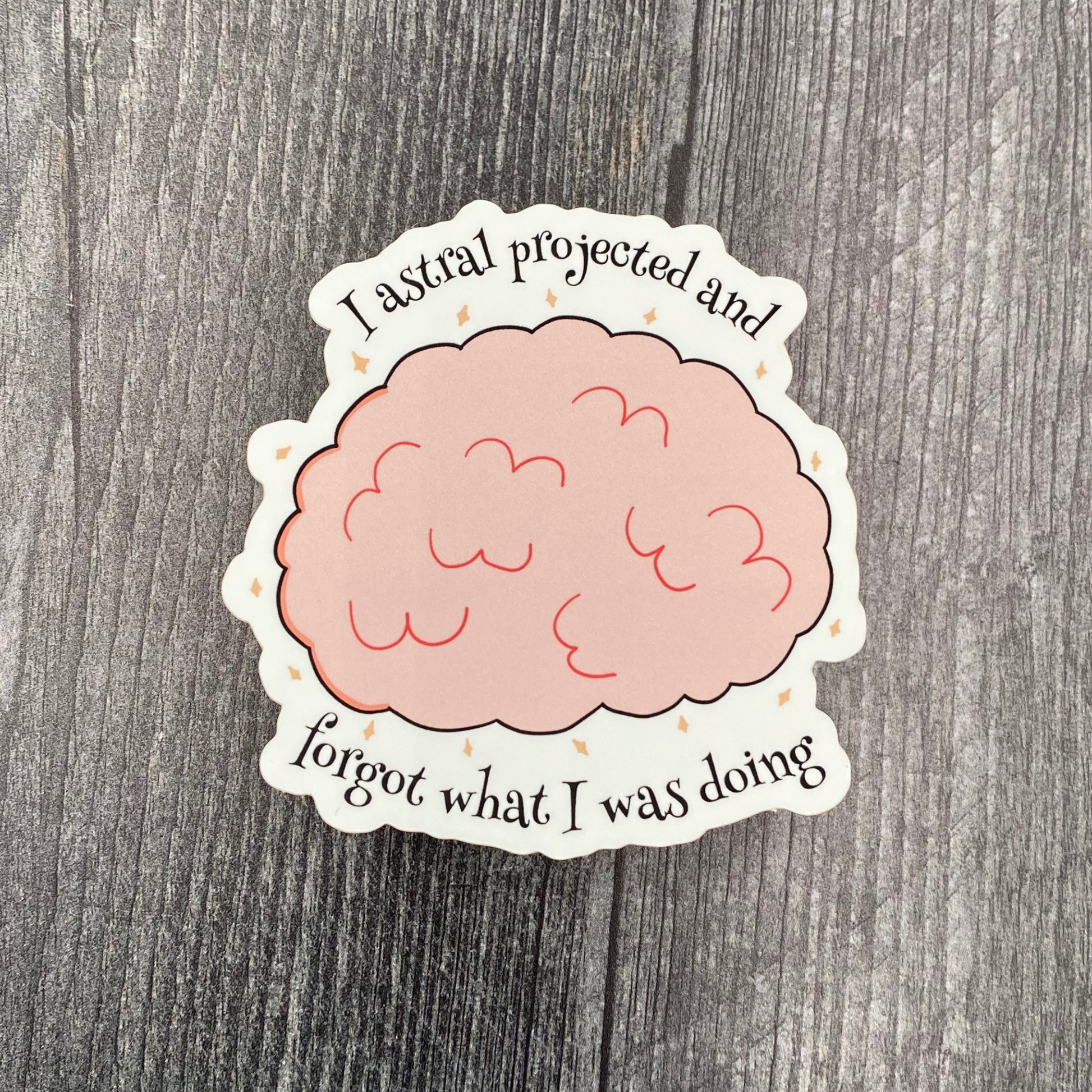 Die-cut vinyl sticker showing a pink brain surrounded by sparkles, with the text "I astral projected and forgot what I was doing," on a gray wood background