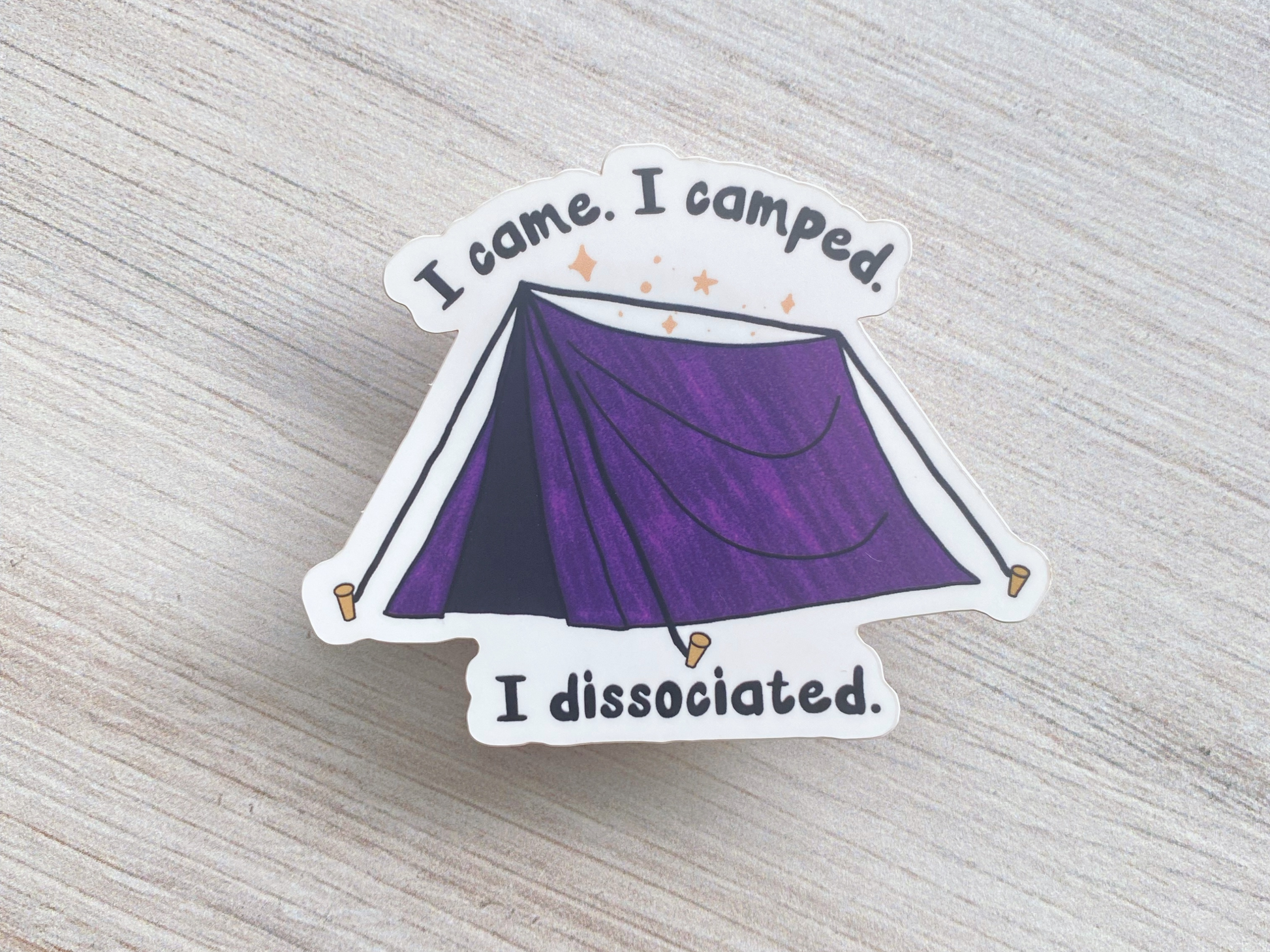Die-cut vinyl sticker showing a purple camping tent under stars, with the text "I came, I camped, I dissociated," on a light wood background
