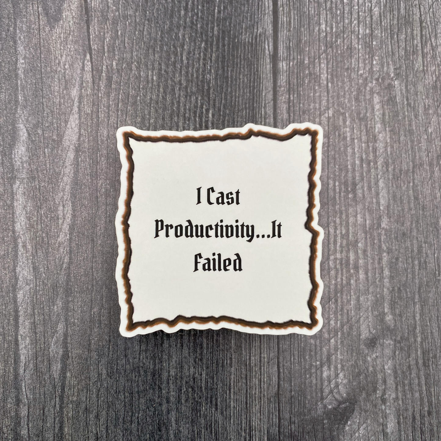 Die-cut vinyl sticker showing a scorched parchment scroll with gothic text reading "I cast productivity, it failed," on a gray wood background