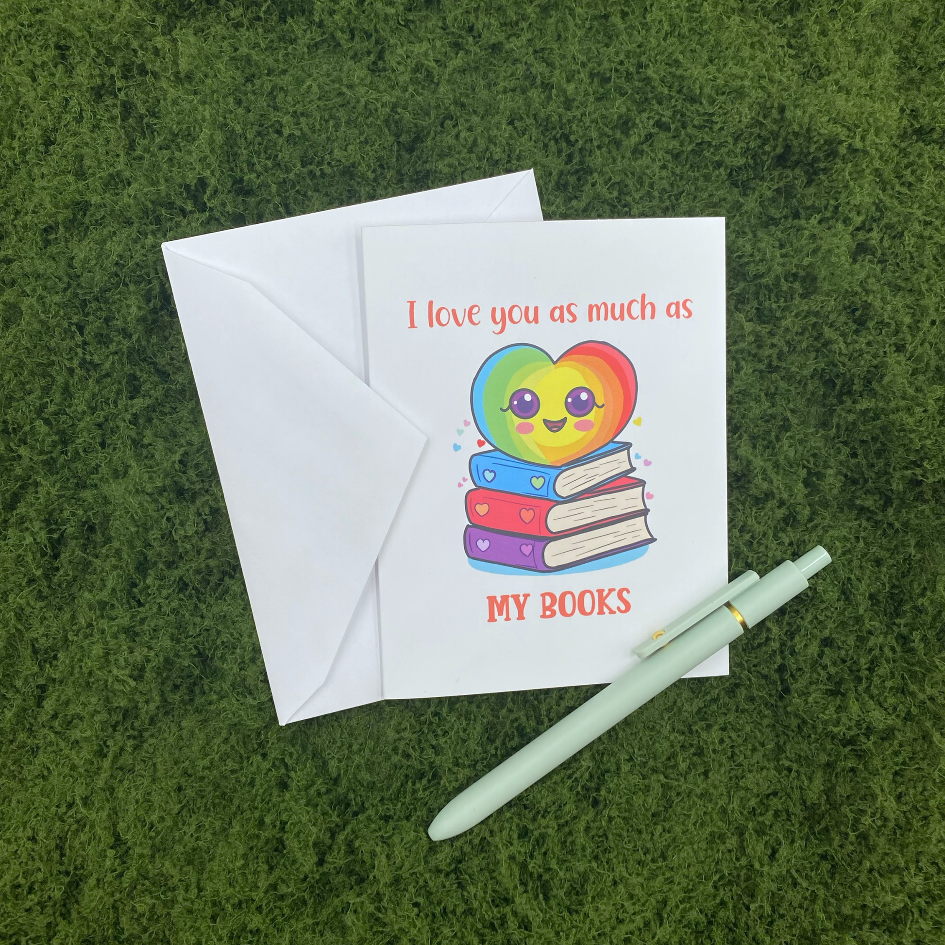 Greeting card that reads "I love you as much as my books" with an illustration of a smiling rainbow heart sitting on a stack of books, propped against its envelope on artificial grass next to a mint green pen.