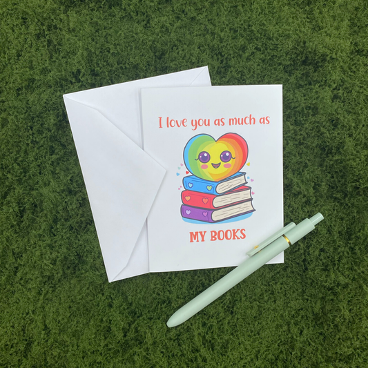 Greeting card that reads "I love you as much as my books" with an illustration of a smiling rainbow heart sitting on a stack of books, propped against its envelope on artificial grass next to a mint green pen.