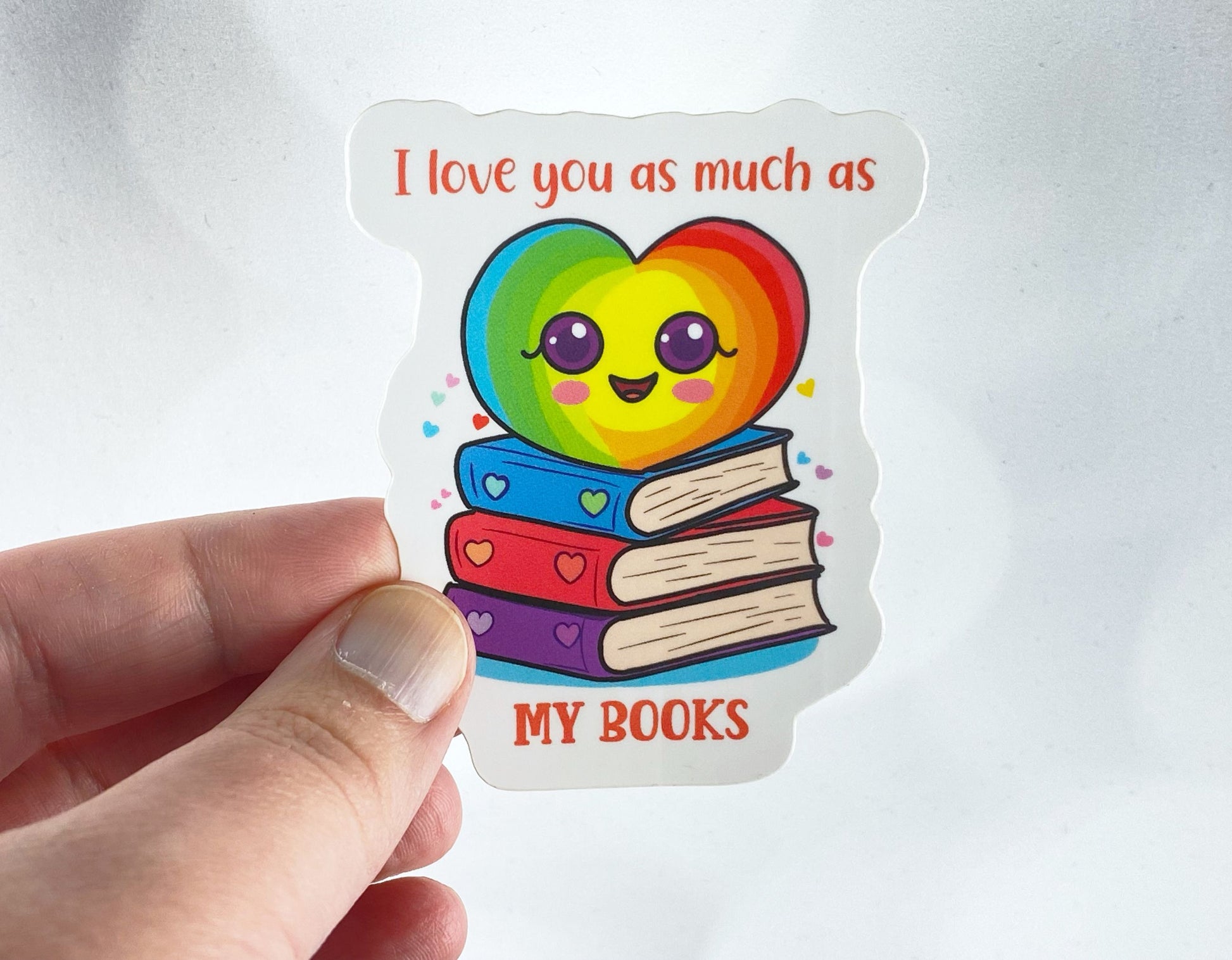 Die-cut vinyl sticker held between fingers, showing a rainbow heart character sitting on a stack of books, above the text "I love you as much as my books," on a white background
