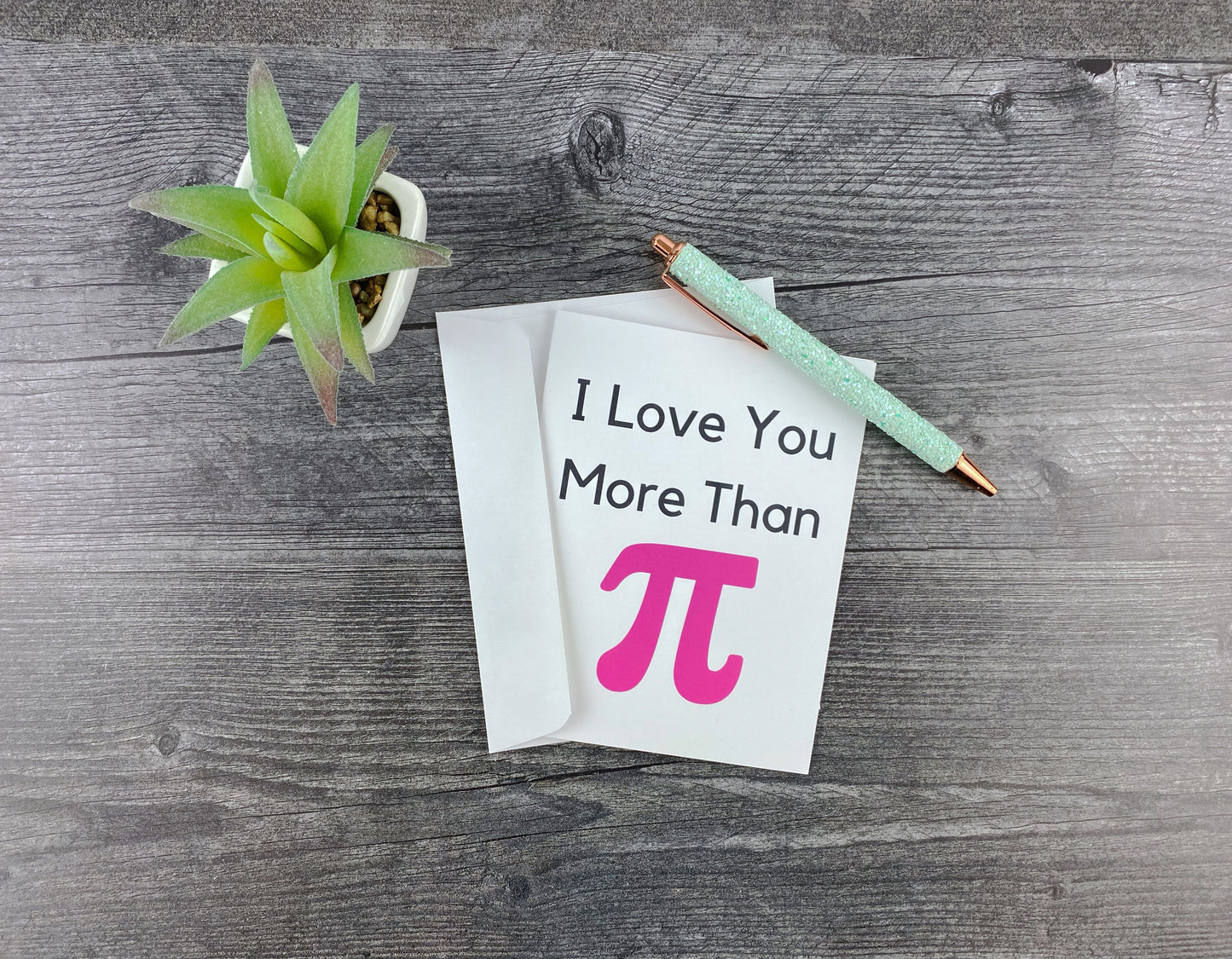 Greeting card and matching envelope, front reading "I love you more than pi" with a large pink pi symbol, resting on a grey wood surface next to a mint glitter pen and small succulent.