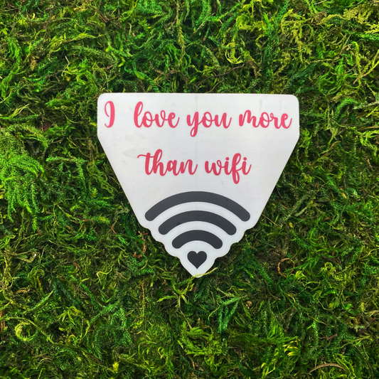 Die-cut matte sticker shaped like a wifi symbol with a small heart at the base, featuring the text "I love you more than wifi" in pink cursive script, displayed on a mossy green surface.