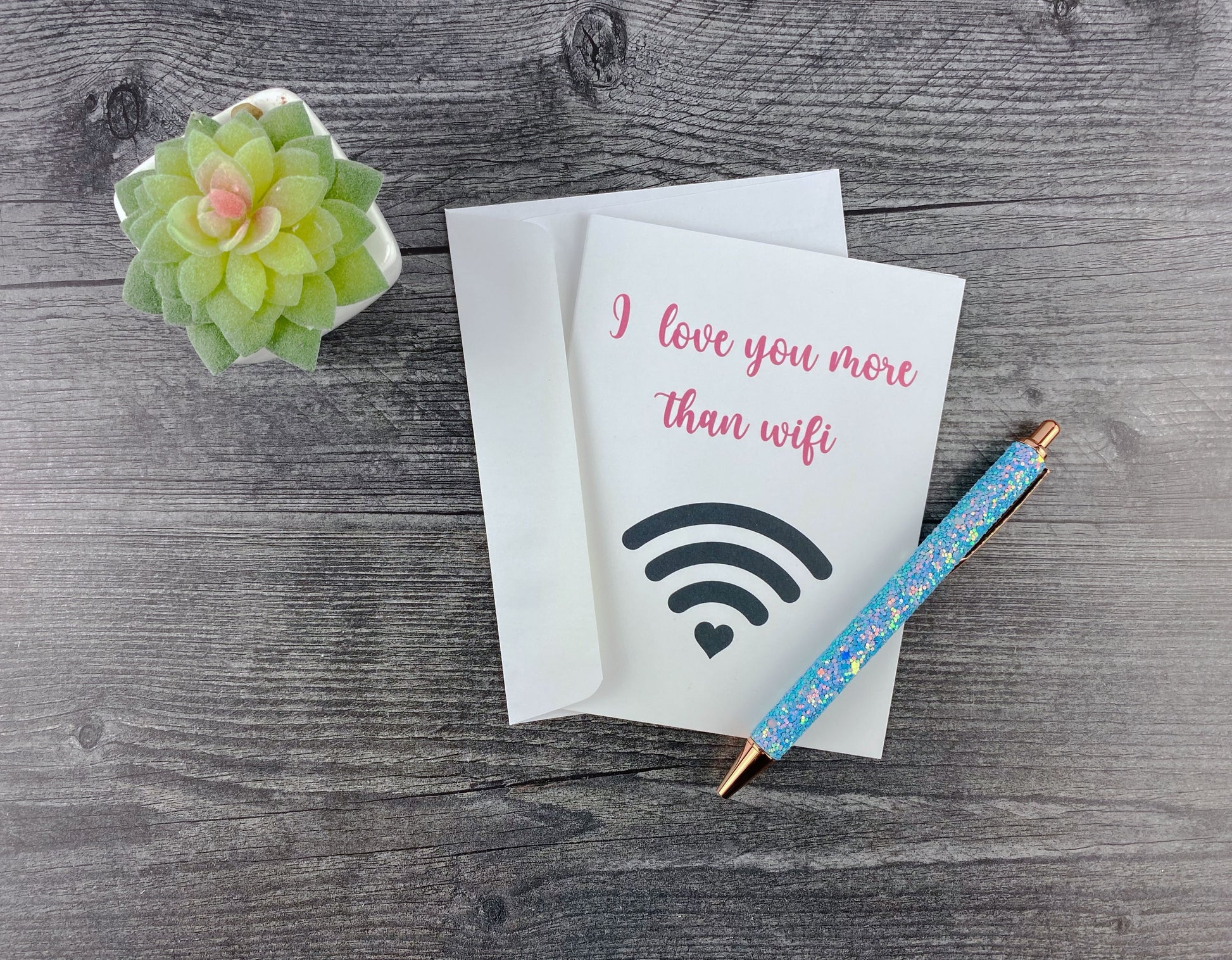Greeting card and matching envelope, front reading "I love you more than wifi" with a wifi signal icon ending in a heart, resting on a grey wood surface next to a blue glitter pen and small succulent.