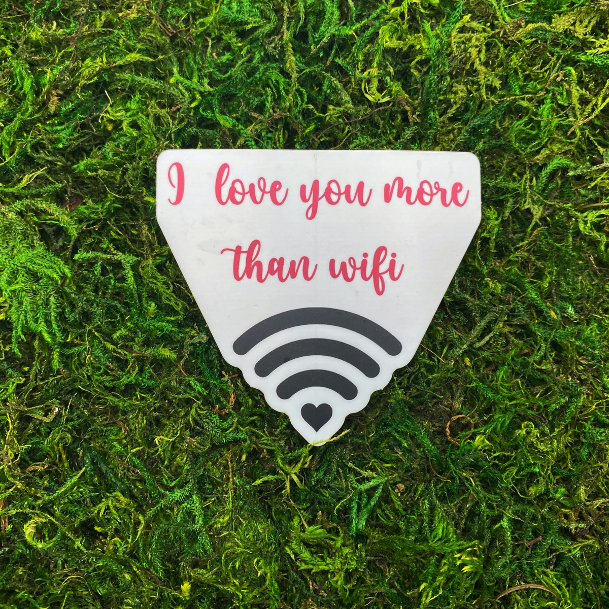 A sticker reading "I love you more than wifi" above a wifi signal icon with a heart at its center, resting on artificial moss.