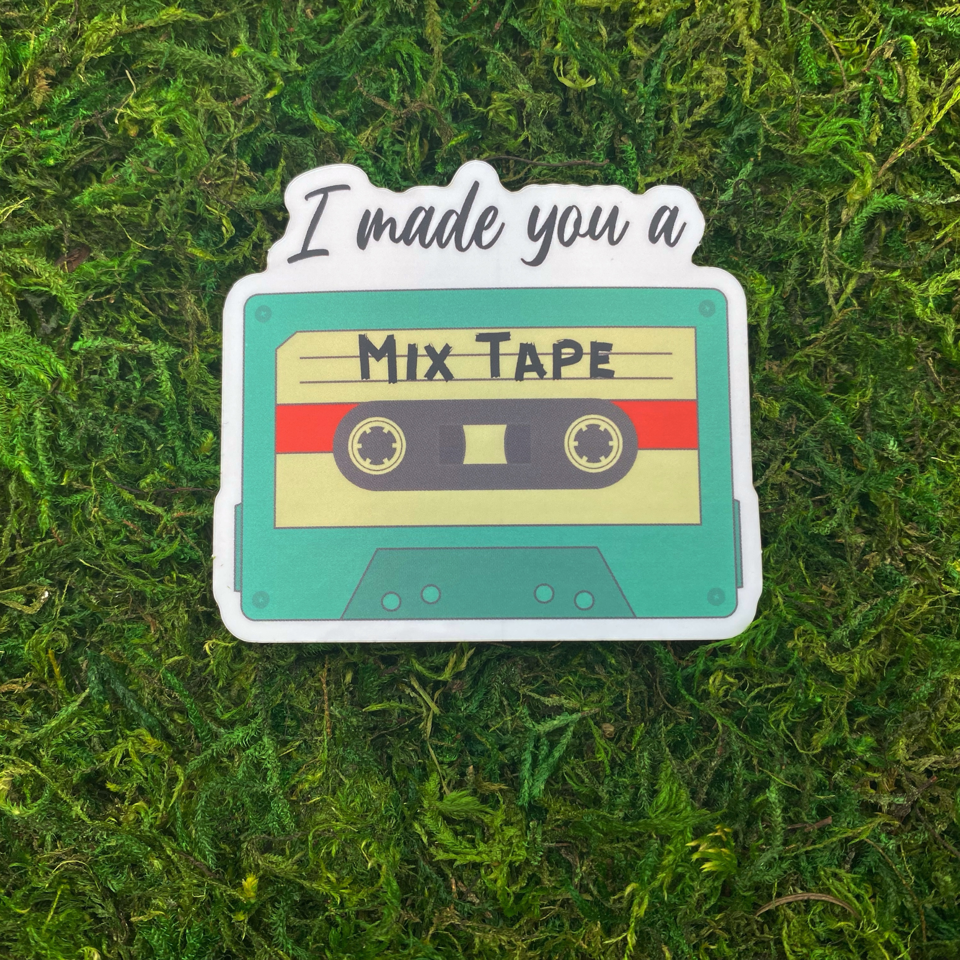 A sticker illustration of a teal cassette tape labeled "Mix Tape" with the text "I made you a" above it, resting on artificial moss.