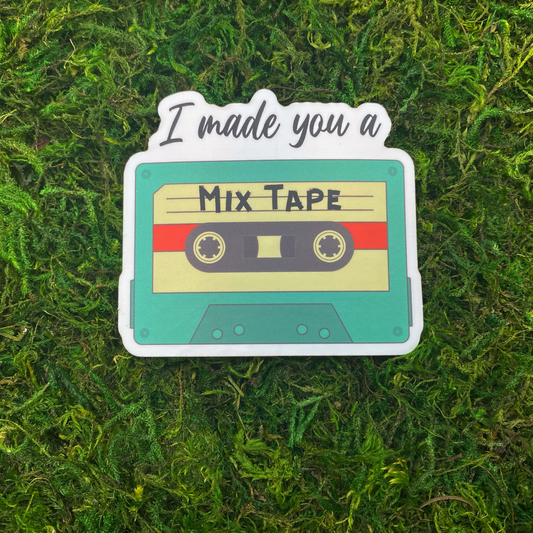 A sticker illustration of a teal cassette tape labeled "Mix Tape" with the text "I made you a" above it, resting on artificial moss.