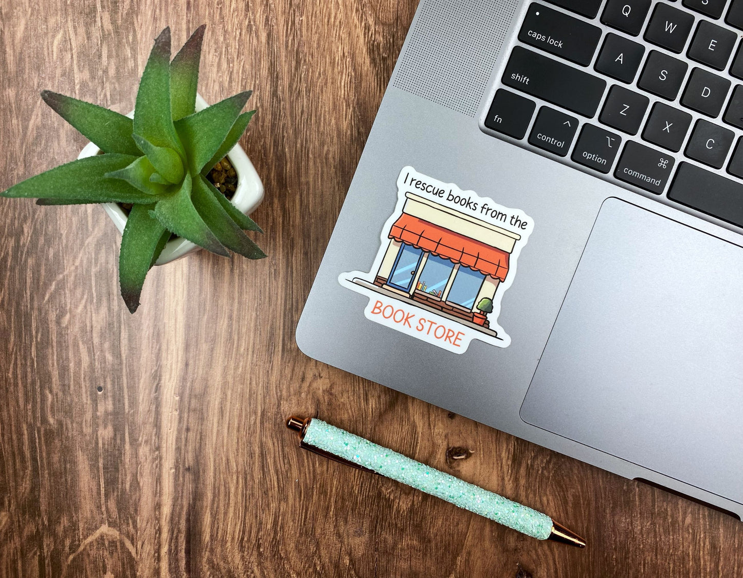 Bookshop storefront sticker applied to the corner of a silver laptop next to a succulent and a glitter pen on a wood desk