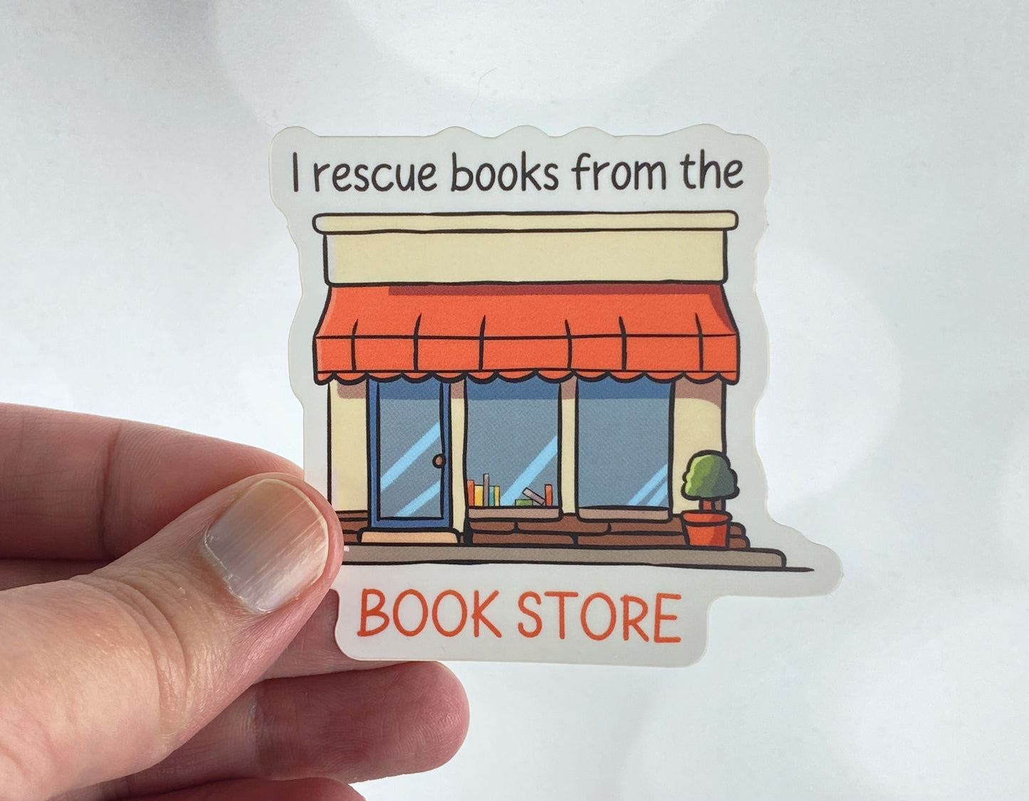 Die-cut vinyl sticker held between fingers, showing a cozy bookshop storefront with an orange awning and books in the window, with the text "I rescue books from the book store," on a white background