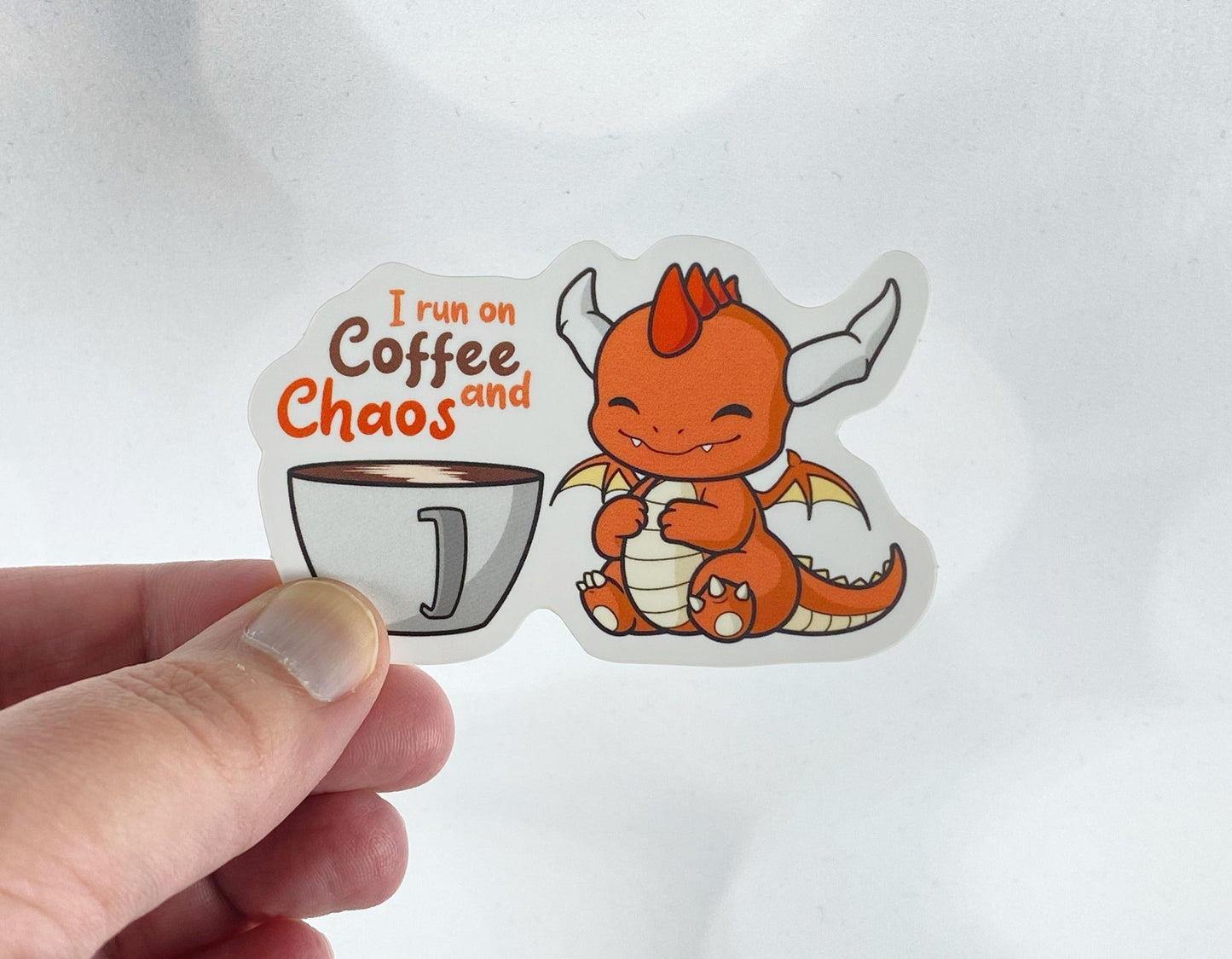 Die-cut vinyl sticker held between fingers, showing an orange dragon sitting next to a cup of coffee, with the text "I run on coffee and chaos," on a white background