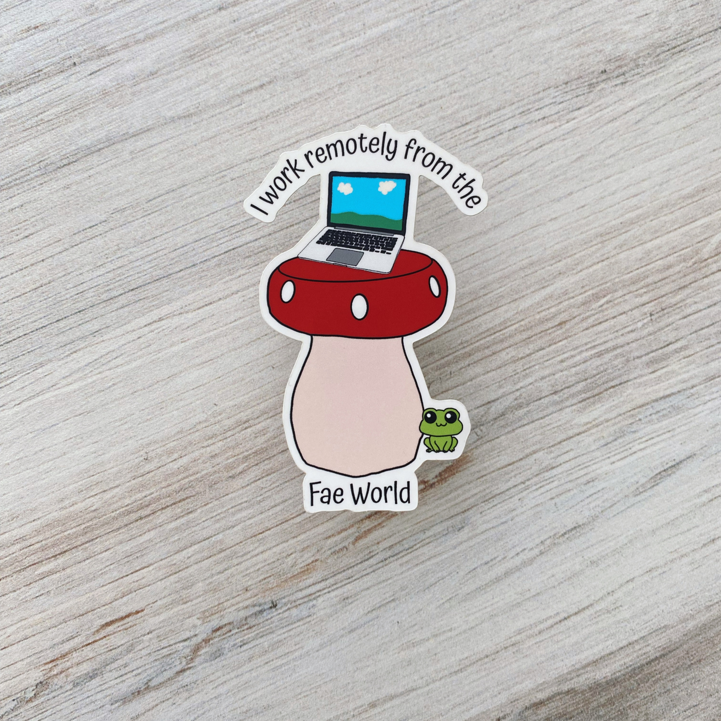 Die-cut vinyl sticker showing a laptop on top of a red mushroom with a green frog nearby, with the text "I work remotely from the Fae World," on a light wood background