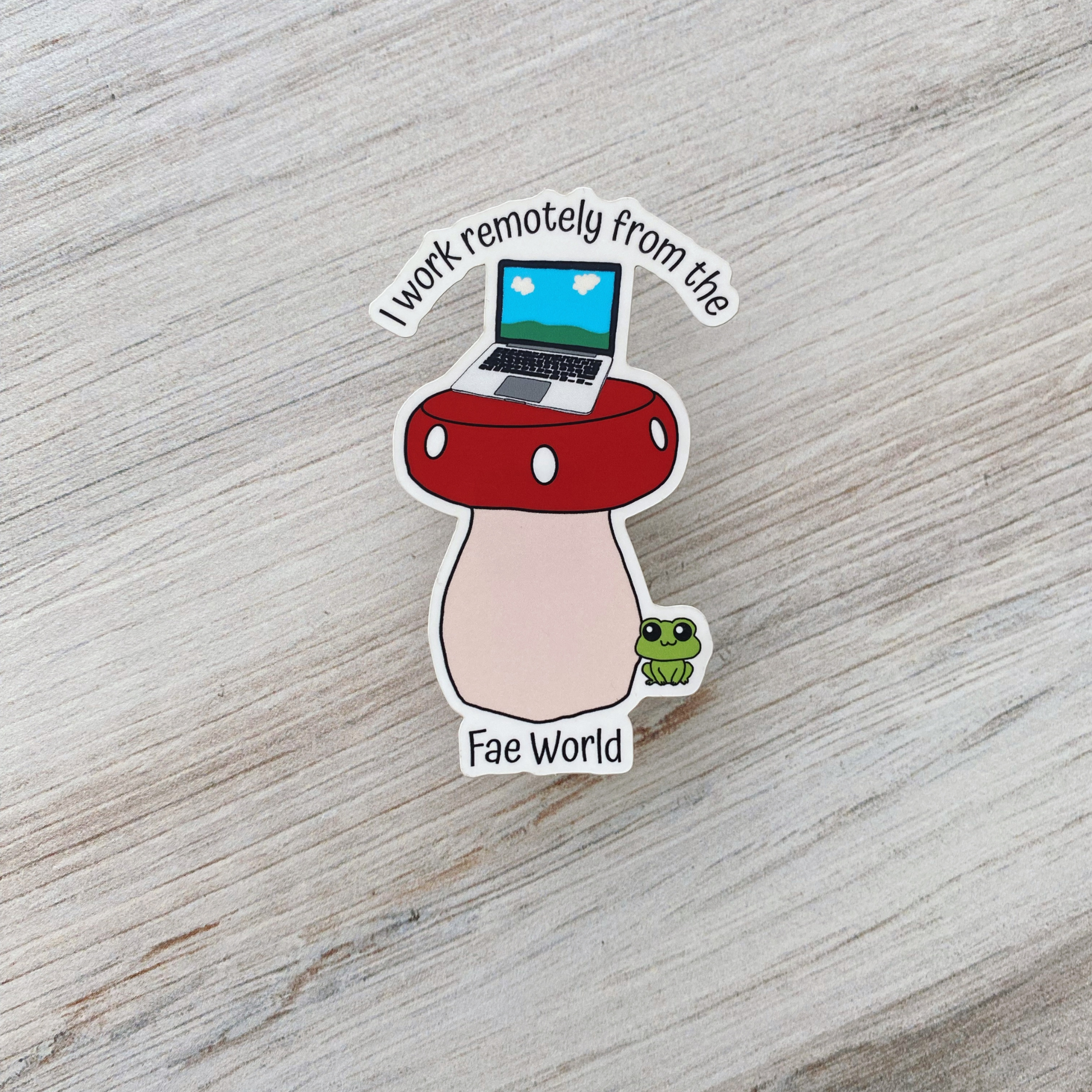Die-cut vinyl sticker showing a laptop on top of a red mushroom with a green frog nearby, with the text "I work remotely from the Fae World," on a light wood background